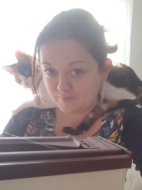 Le Chat des Aiguilles Couturière en train de travailler sur sa machine à coudre avec un chat tricolore allongé sur ses épaules