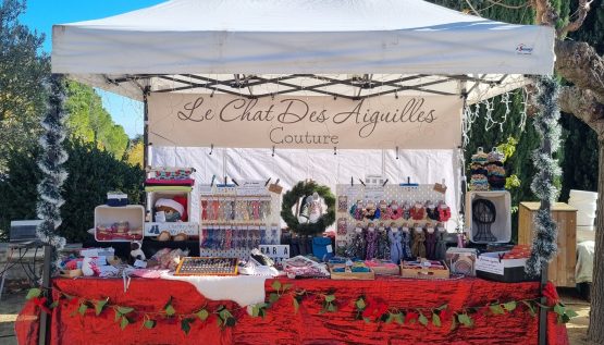 Stand de marché de Noël par un beau soleil. Nappe rouge brillante, guirlandes de Noël, décoration dans le thème des fêtes. Sur le stand, des accessoires cousus main. Banderole en fond de barnum avec le nom de la marque, le Chat des Aiguilles.
