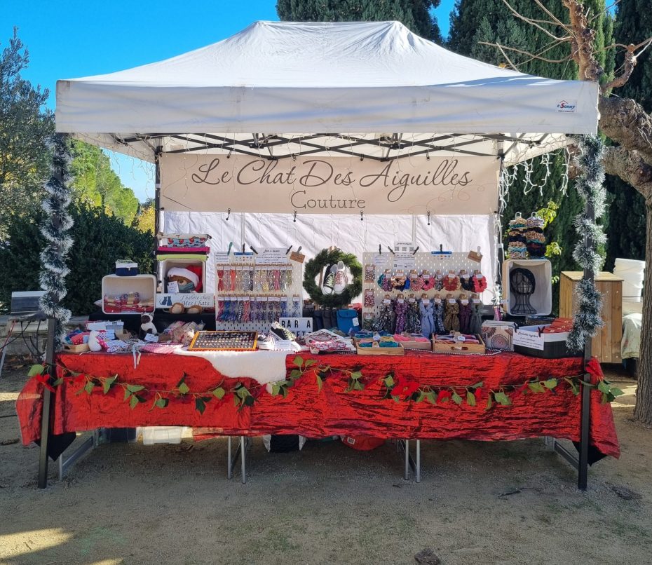 Stand de marché de Noël par un beau soleil. Nappe rouge brillante, guirlandes de Noël, décoration dans le thème des fêtes. Sur le stand, des accessoires cousus main. Banderole en fond de barnum avec le nom de la marque, le Chat des Aiguilles.